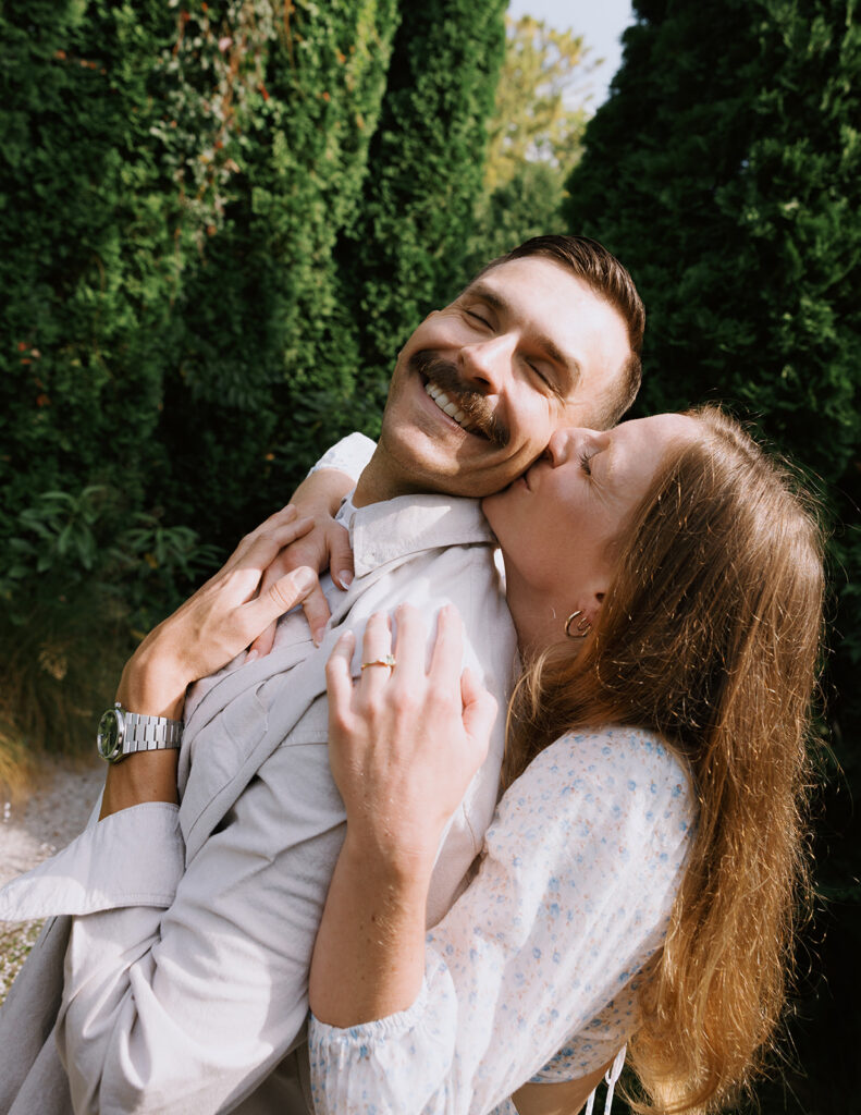 Close-up of a joyful couple in a lush garden, with the woman hugging the man from behind and kissing his cheek as he smiles with his eyes closed.